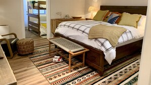 Quarto com cama de madeira, um banquinho, mesa de cabeceira, abajur e tapete estampado.