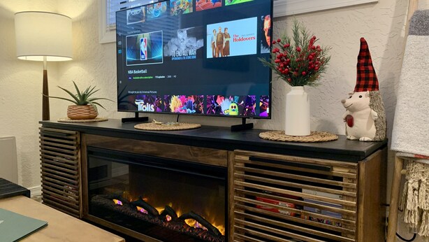 Uma sala de estar com televisão de tela plana exibindo diversos canais, lareira, pequena mesa com um laptop e objetos decorativos, incluindo uma planta e um pequeno ouriço de brinquedo.