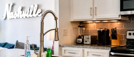 A kitchen with a stainless steel faucet, a countertop with a bottle of hand soap, a green bottle, and a sponge.