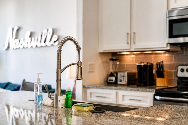 A kitchen with a stainless steel faucet, a countertop with a bottle of hand soap, a green bottle, and a sponge.