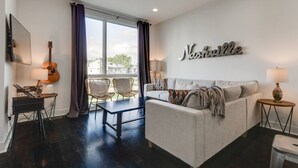 A living room with a grey sofa, two armchairs, a coffee table, a guitar, a TV, and a large window with curtains. - King-bed townhome near The Gulch | 2-Car Garage (Nashville)