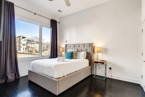 A bedroom with a large bed, two bedside lamps, a window with curtains, and a view of buildings outside.