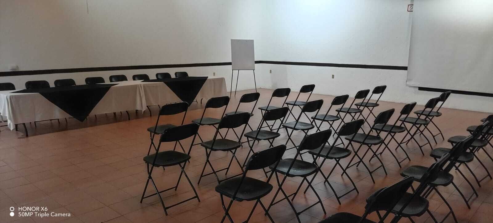 Uma sala de conferências com fileiras de cadeiras pretas dobráveis, um quadro branco e uma mesa com toalha preta.