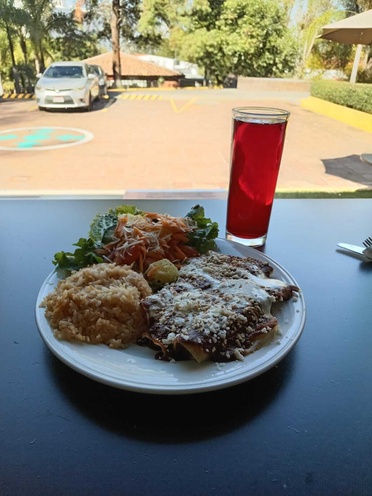Un plato de comida con arroz, un guiso de carne y verduras, y un vaso de bebida roja sobre una mesa con vistas a la calle y coches aparcados.