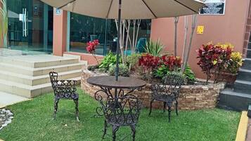 Een terras met een tafel, stoelen, een parasol, en een kleine tuin met planten en een bank.