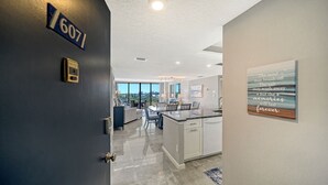 A modern kitchen with a central island, a dining area with a table and chairs, and a view of the outdoors through large windows. - The Anchorage - Unit 607 (Siesta Key)