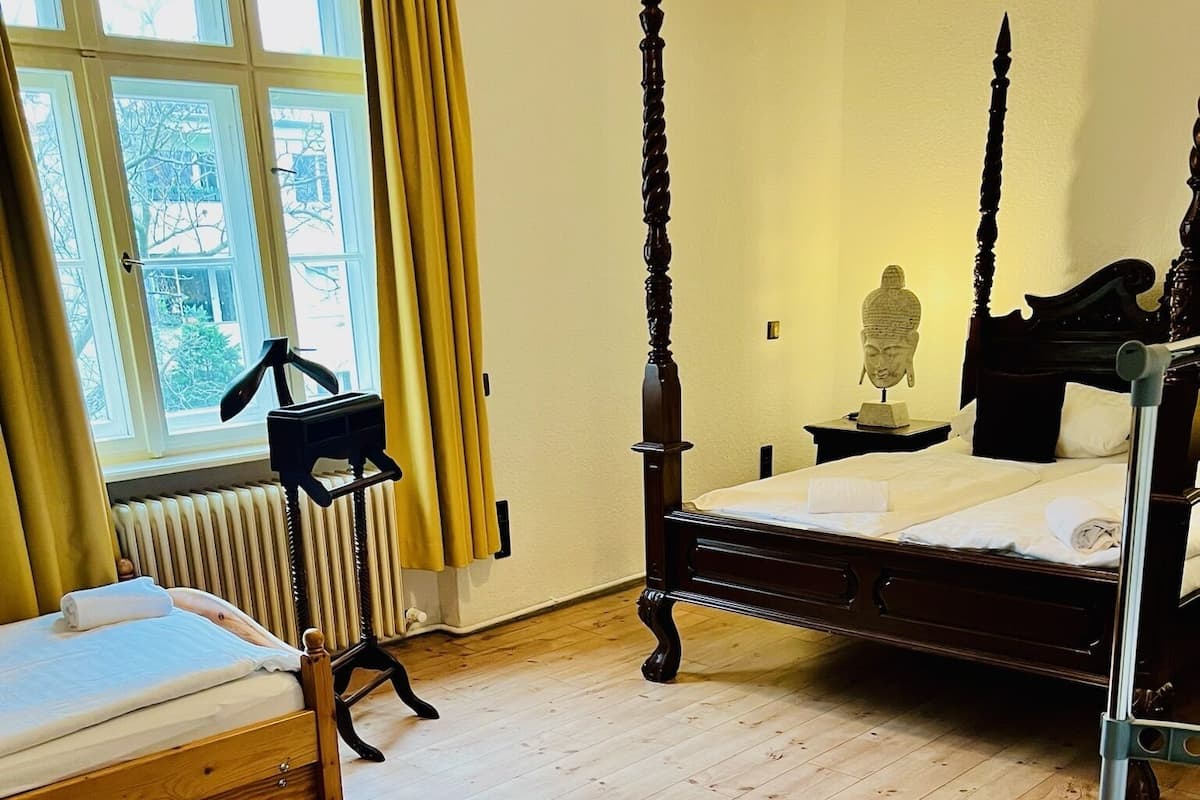 Una habitación con piso de madera, una cama con cabecera, una mesita de noche con lámpara, una ventana con cortinas y un radiador.
