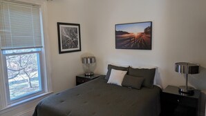 Un dormitorio con una cama, dos mesitas de noche, un ventanal y dos cuadros enmarcados en la pared.