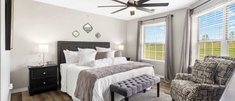 A bedroom with a bed, bedside tables, a bench, a chair, a ceiling fan, and large windows with curtains.