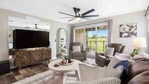 A spacious living room with a flat-screen TV, a ceiling fan, and a large window offering a view of the outdoors. - Catch the Sunrise - Branson Mo - Holiday Hills - 2bd Condo! (Branson)
