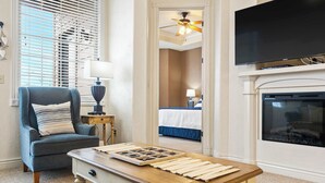 A hotel room with a wooden coffee table, a blue armchair, a bed with a blue bedspread, a television mounted above a fireplace, and a ceiling fan. - Cliffside Lake Retreat - 2 Bdr, Walk to Table Rock (Branson)