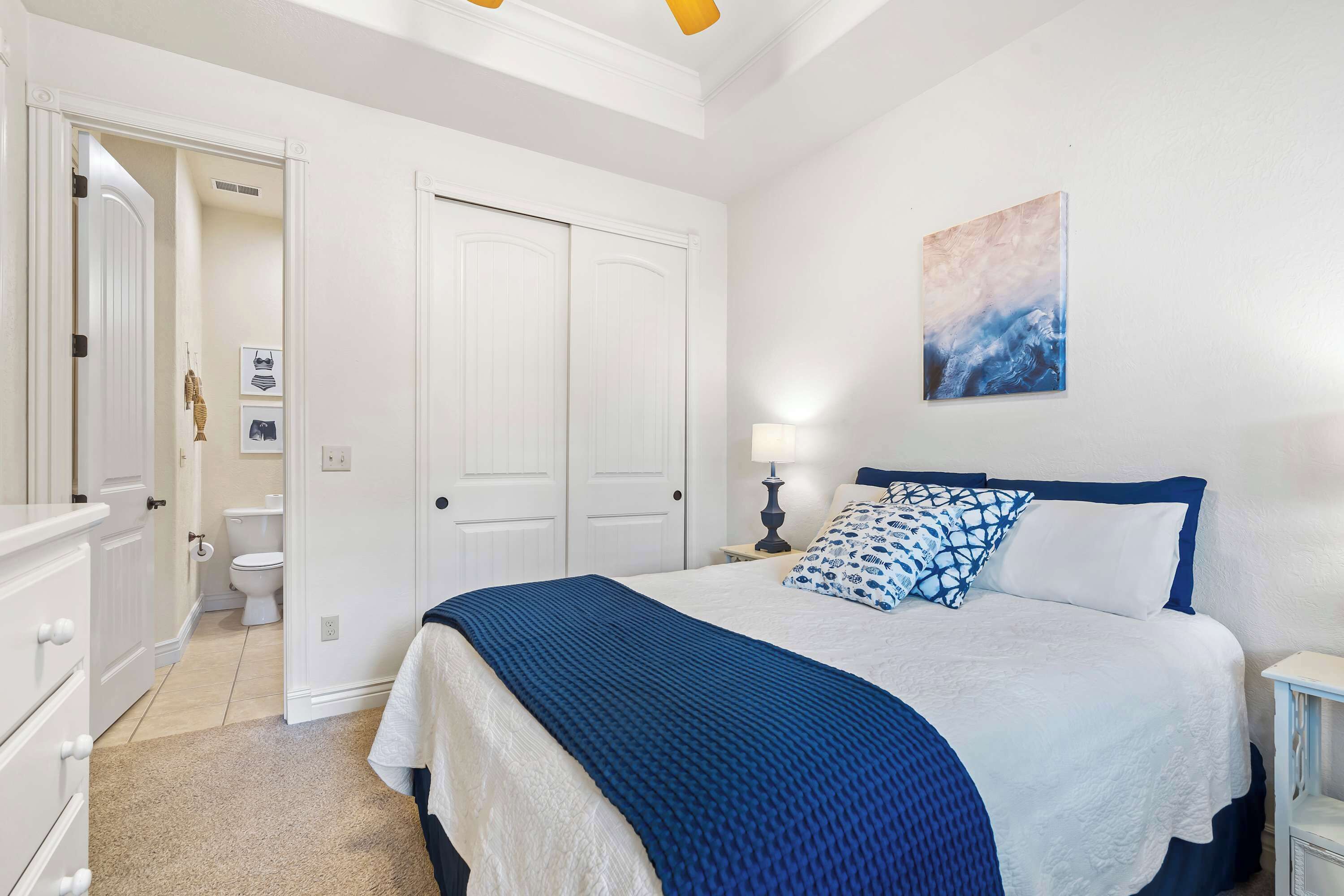 A bedroom with a bed, a nightstand, a lamp, a painting, and a door leading to a bathroom.