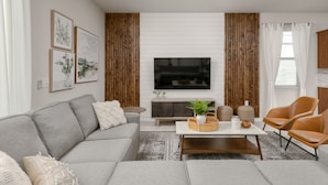 Un salon moderne avec un canapé gris, une table basse et un téléviseur fixé à un mur blanc. De chaque côté du téléviseur, on trouve deux panneaux en bois, et une fenêtre est agrémentée de rideaux.
