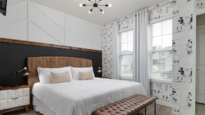 Une chambre avec un grand lit, une tête de lit en bois, un banc, une table de chevet et un papier peint à l’effigie de Mickey Mouse.