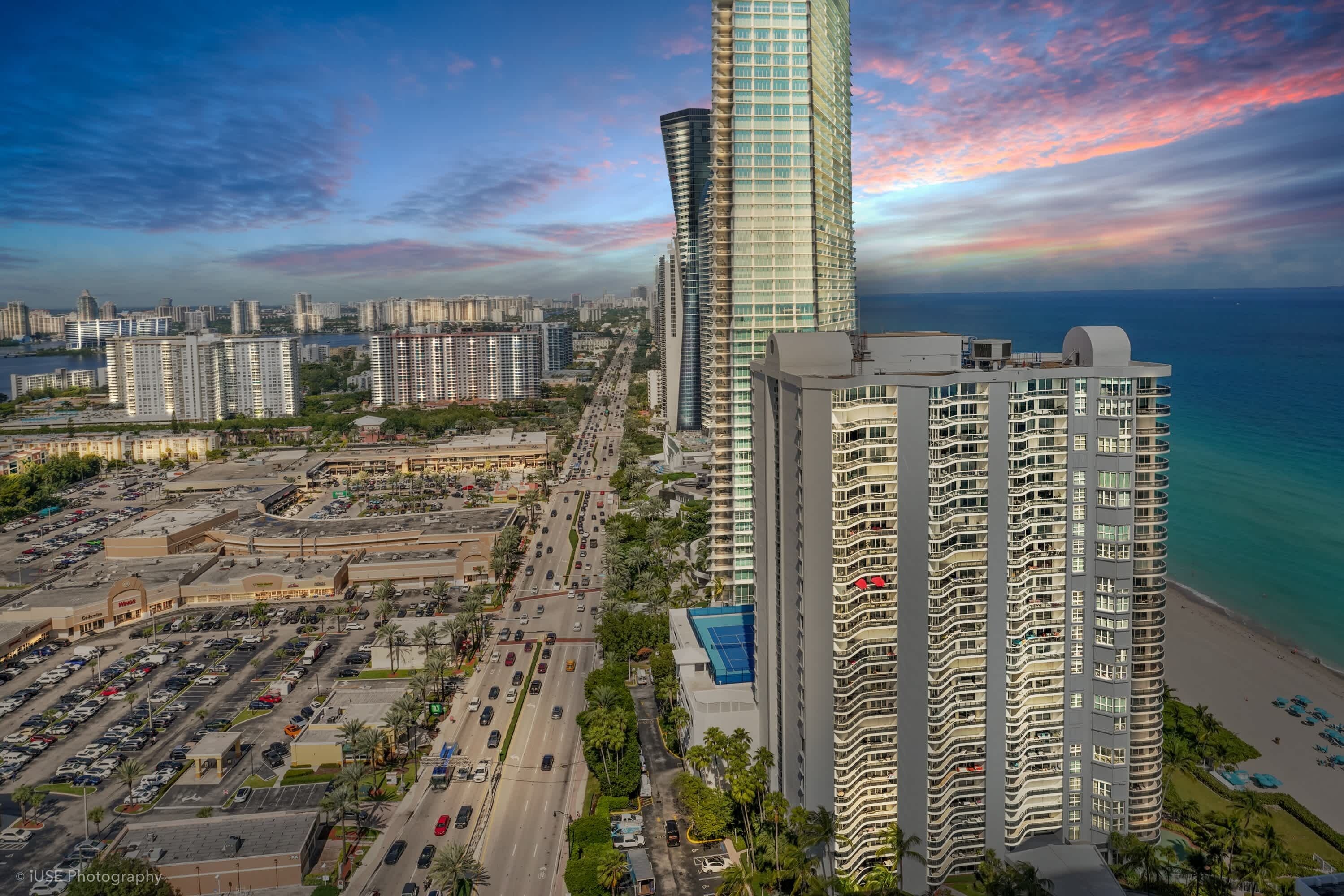A high-rise building with a curved facade, adjacent to a beachfront and a parking lot.