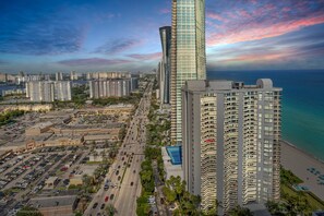 A high-rise building with a curved facade, adjacent to a beachfront and a parking lot.