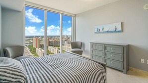 Ein modernes Schlafzimmer mit einem großen Fenster, einem Bett mit gestreifter Bettwäsche, einer grauen Kommode und einem Bild von Segelbooten.