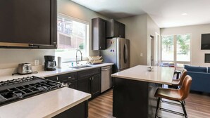 A modern kitchen with dark cabinets, a stainless steel refrigerator, a coffee maker, and a toaster. There is a bar area with a chair and a large window. - Granby Colorful Adventures Breathtaking Views! (Granby)