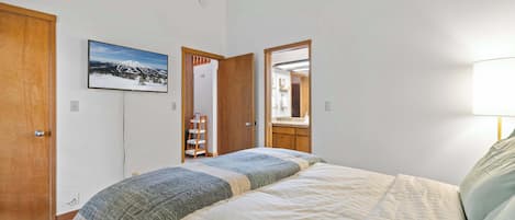 A bedroom with a bed, a wooden door, a framed picture on the wall, and a mirror reflecting a bathroom.