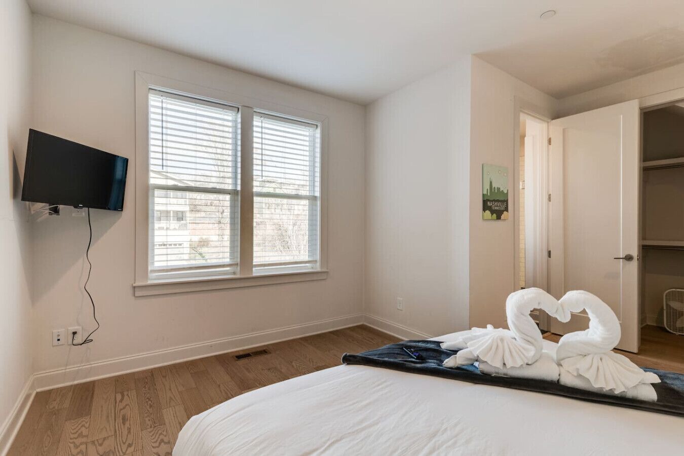 A bedroom with a bed, a TV mounted on the wall, and a window with blinds.