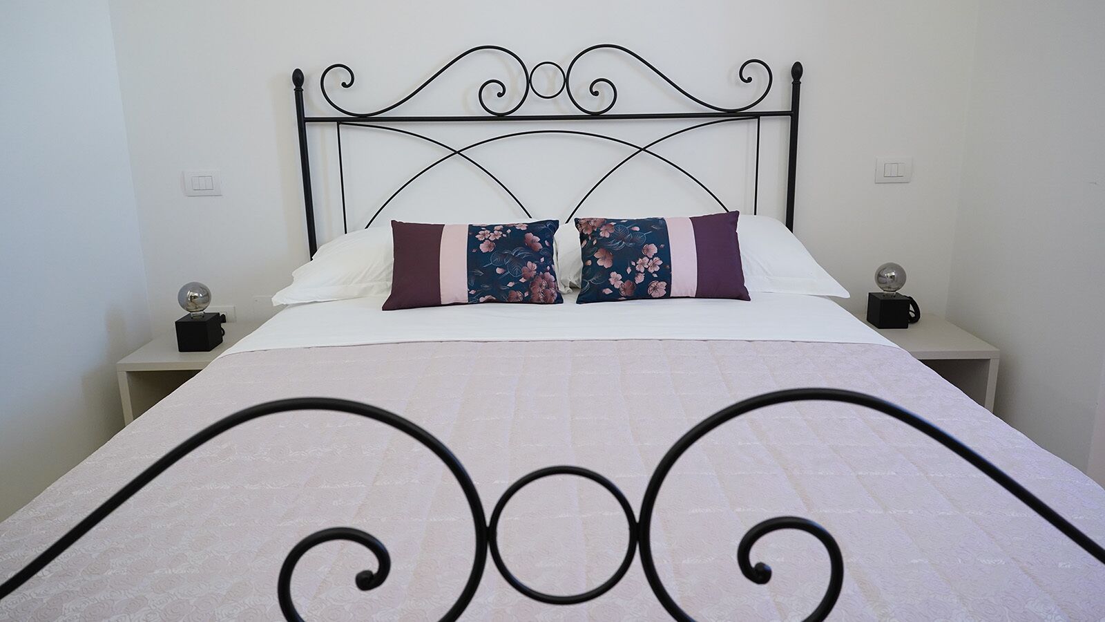 Un lit bien fait, agrémenté d’un coussin à motifs floraux, d’un coussin uni et d’une tête de lit décorative. Deux tables de chevet, chacune surmontée d’une lampe et d’un petit objet décoratif, se trouvent de part et d’autre du lit.