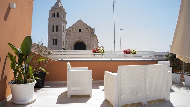Una terraza en la azotea con mobiliario de mimbre blanco, plantas en macetas y vistas a la torre de una iglesia histórica.