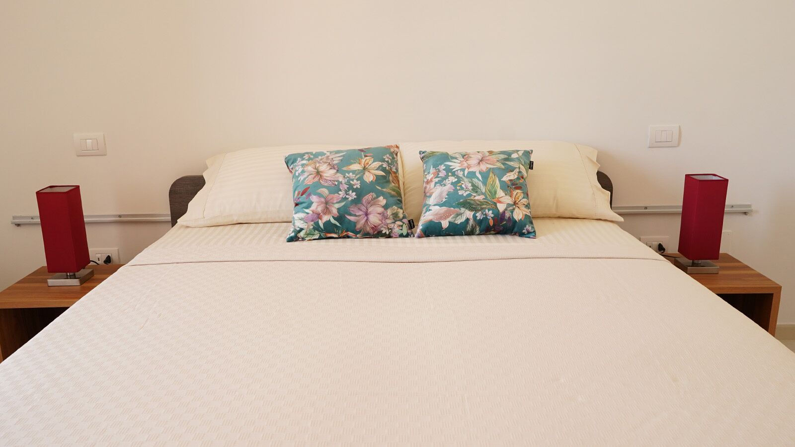 Ein Bett mit zwei blumigen Kissen, einer weißen Bettdecke und zwei Nachttischen mit roten Lampen.