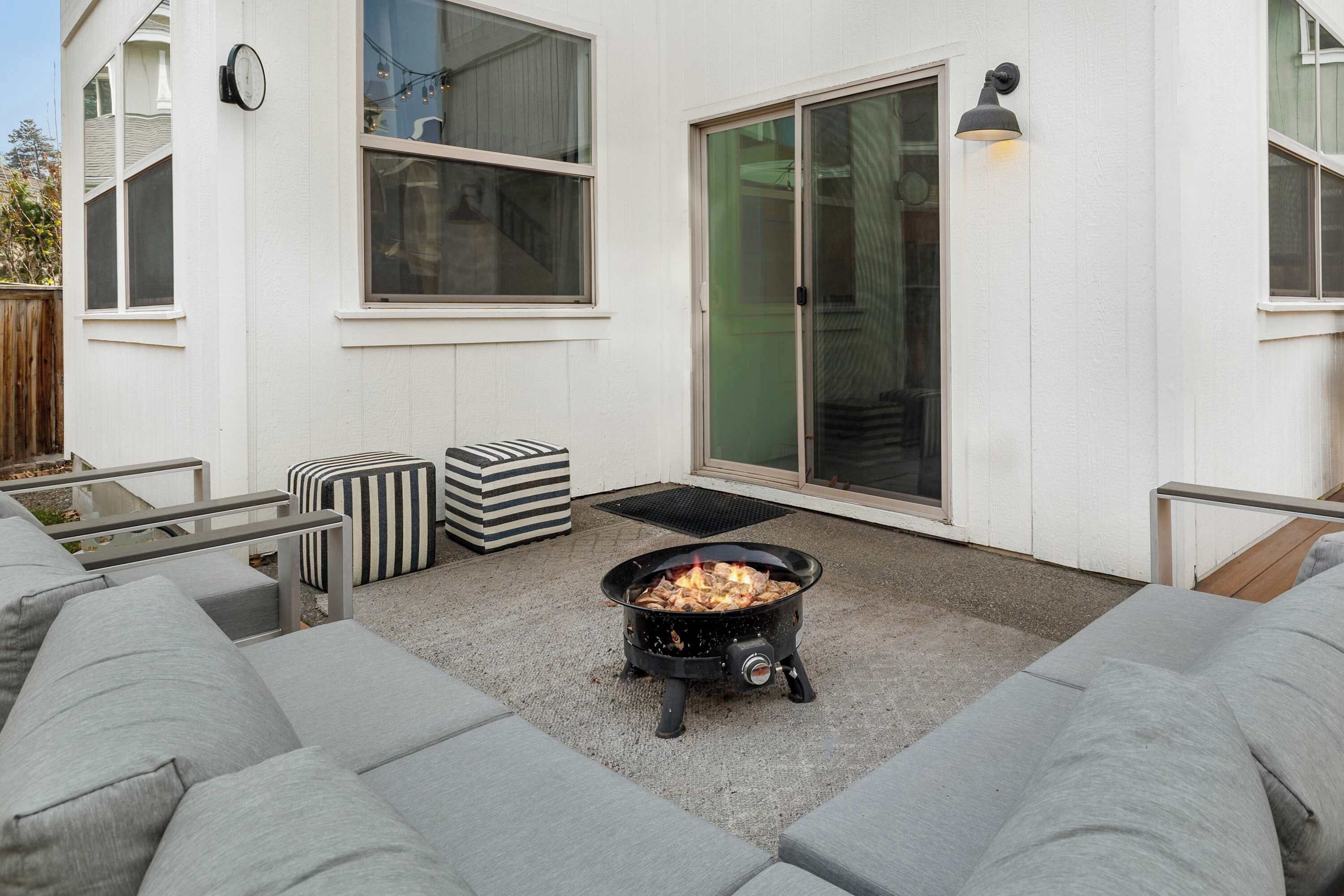 A patio area with a fire pit, grey outdoor furniture, and a white building with large windows and a sliding glass door.