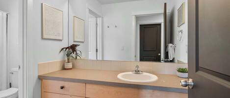 A bathroom with a wooden vanity, a sink, and a mirror with lights. There are framed pictures on the wall and a plant on the vanity.