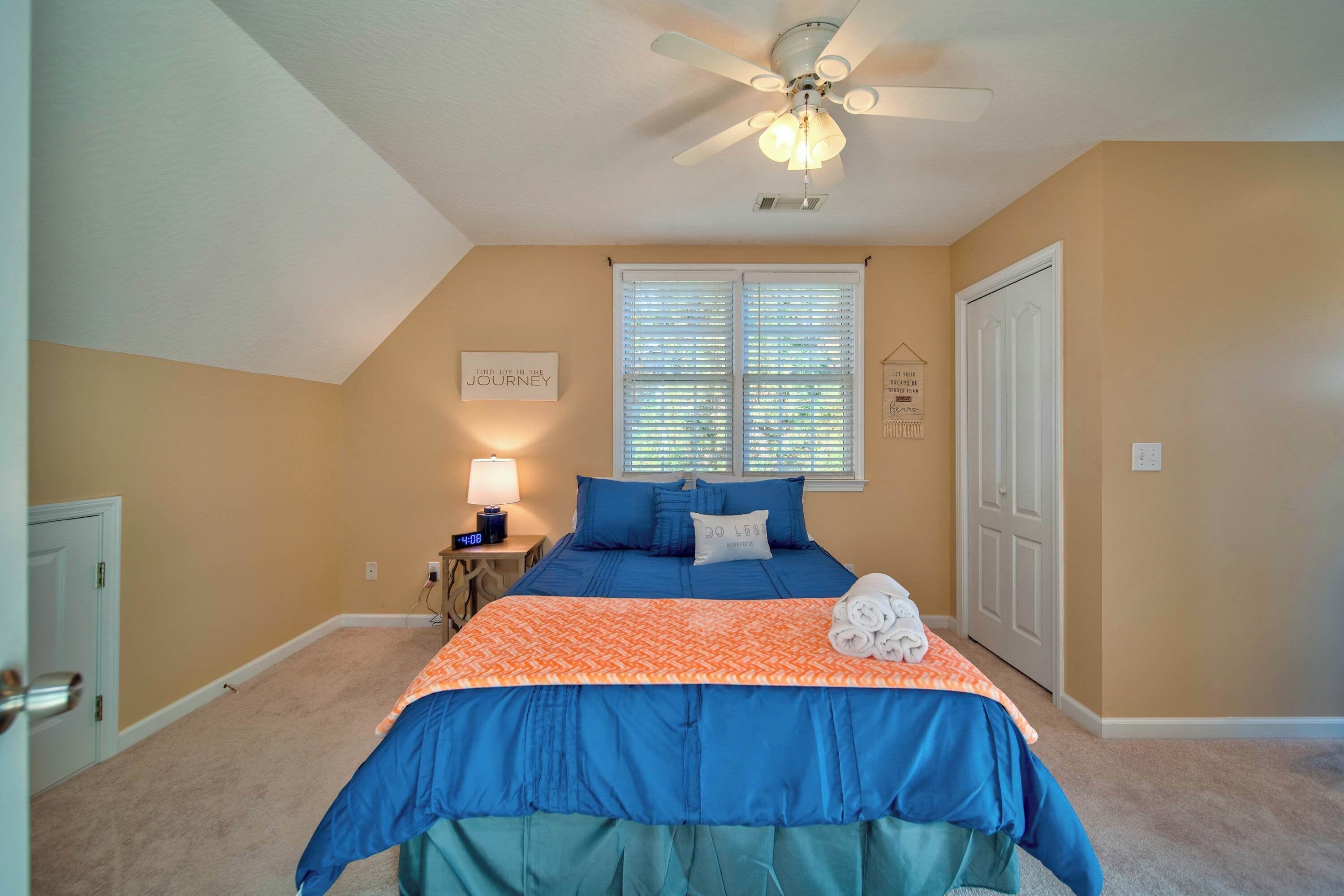 A bedroom with a bed, a ceiling fan, a window with blinds, a nightstand with a lamp, and a door.