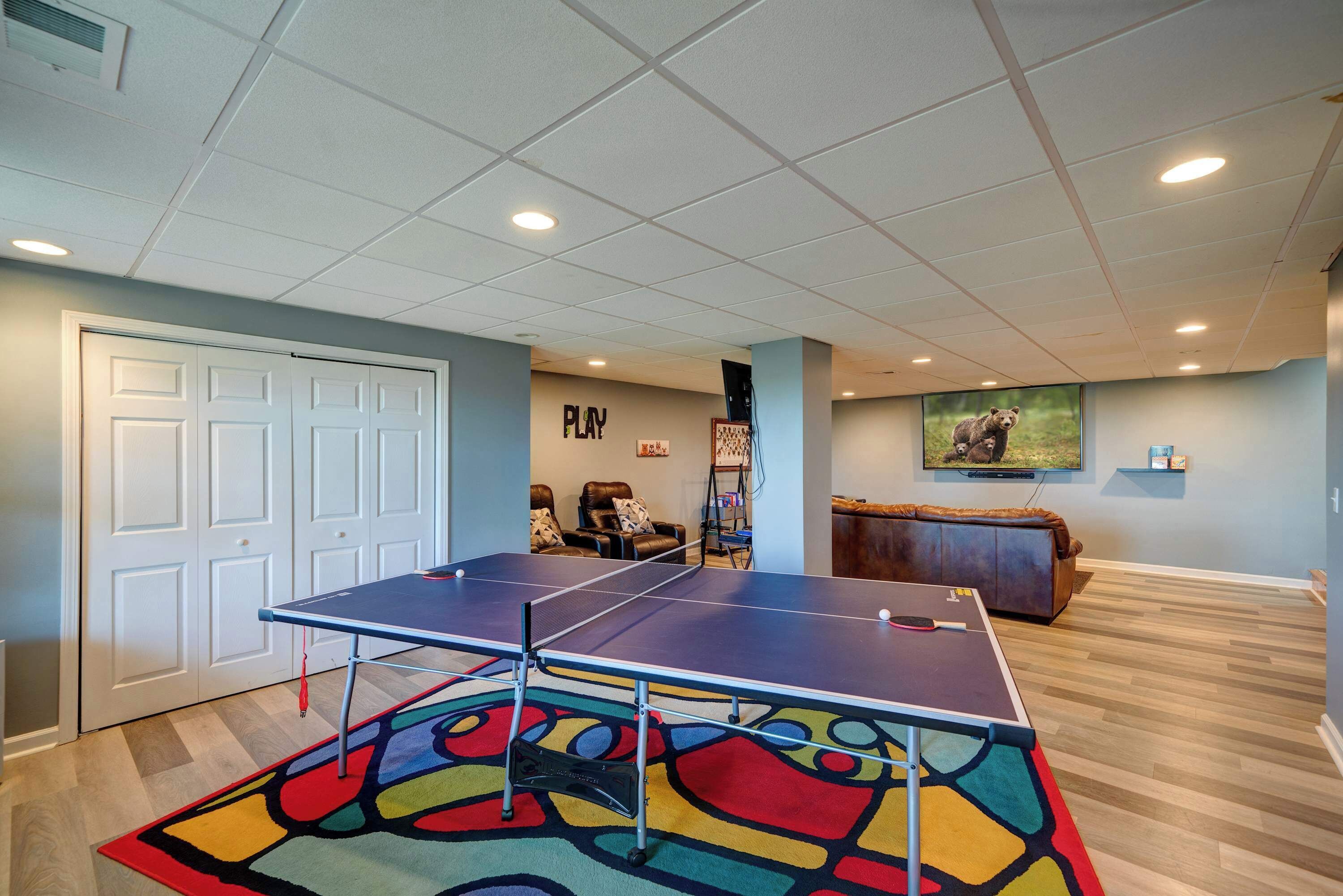A ping pong table in a room with a colorful rug, a couch, and a wall with a bear picture.