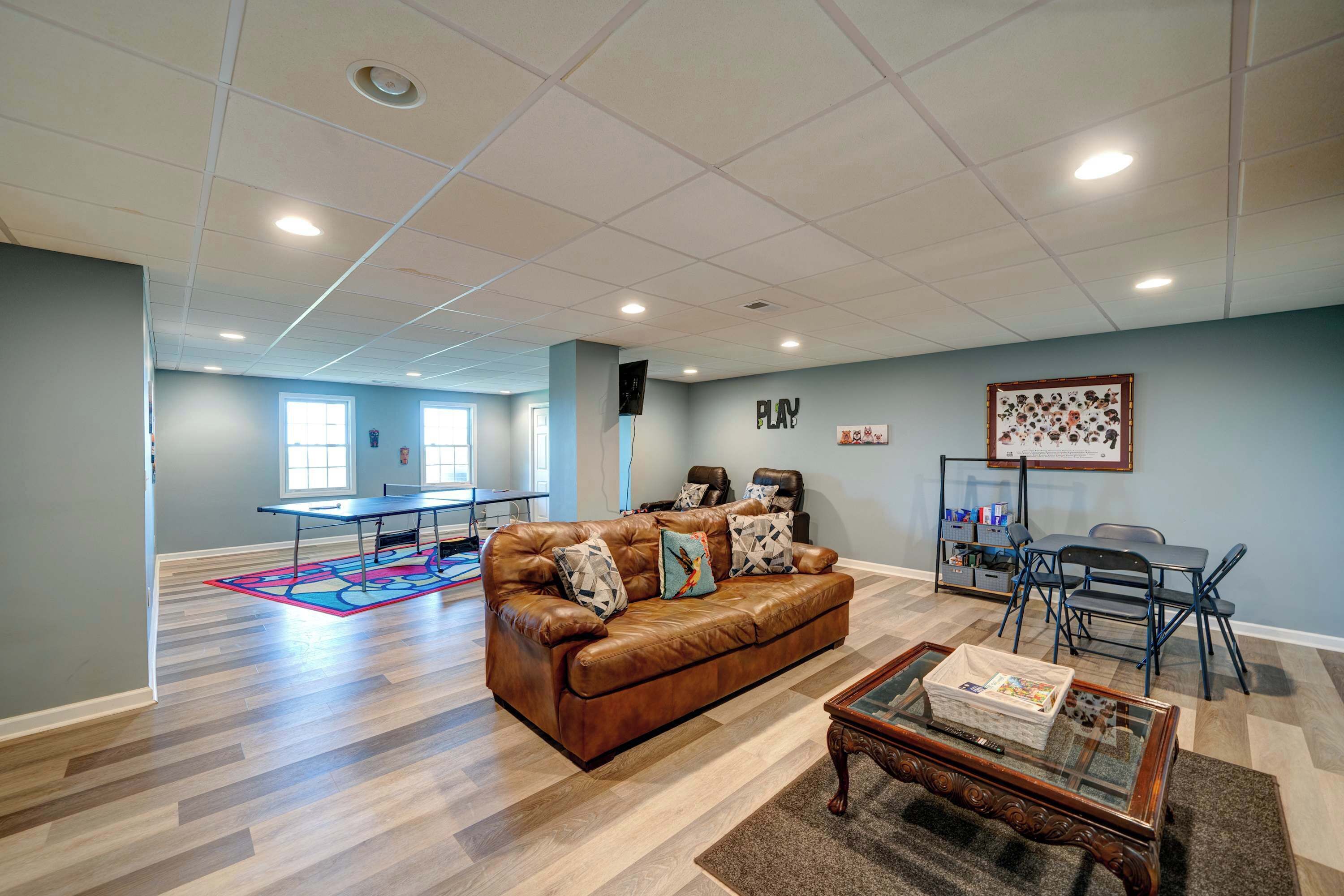 A spacious living room with a brown leather sofa, a glass coffee table, and a ping pong table in the background.