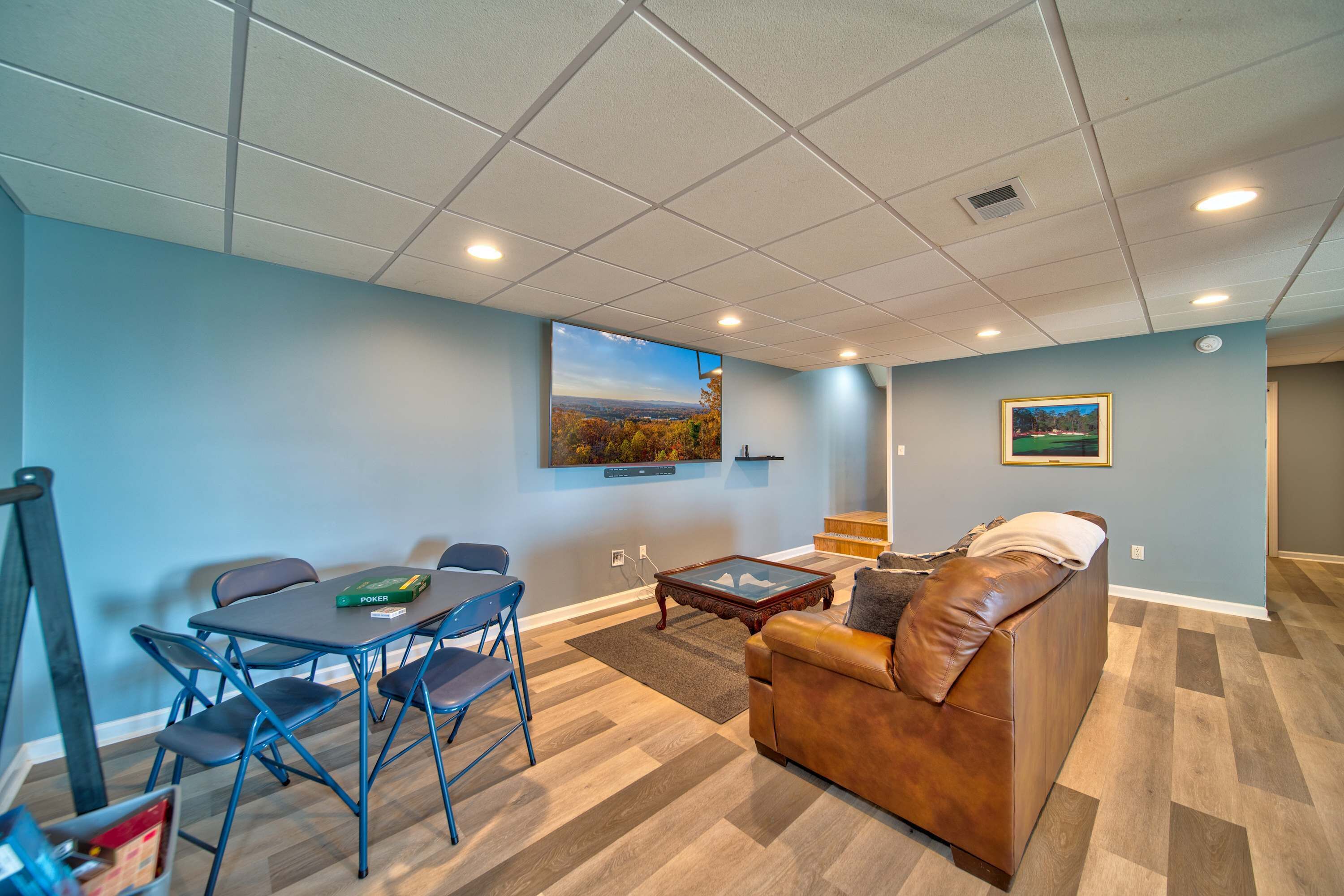 A living room with a brown leather couch, a dining table with chairs, a coffee table, and a painting on the wall.