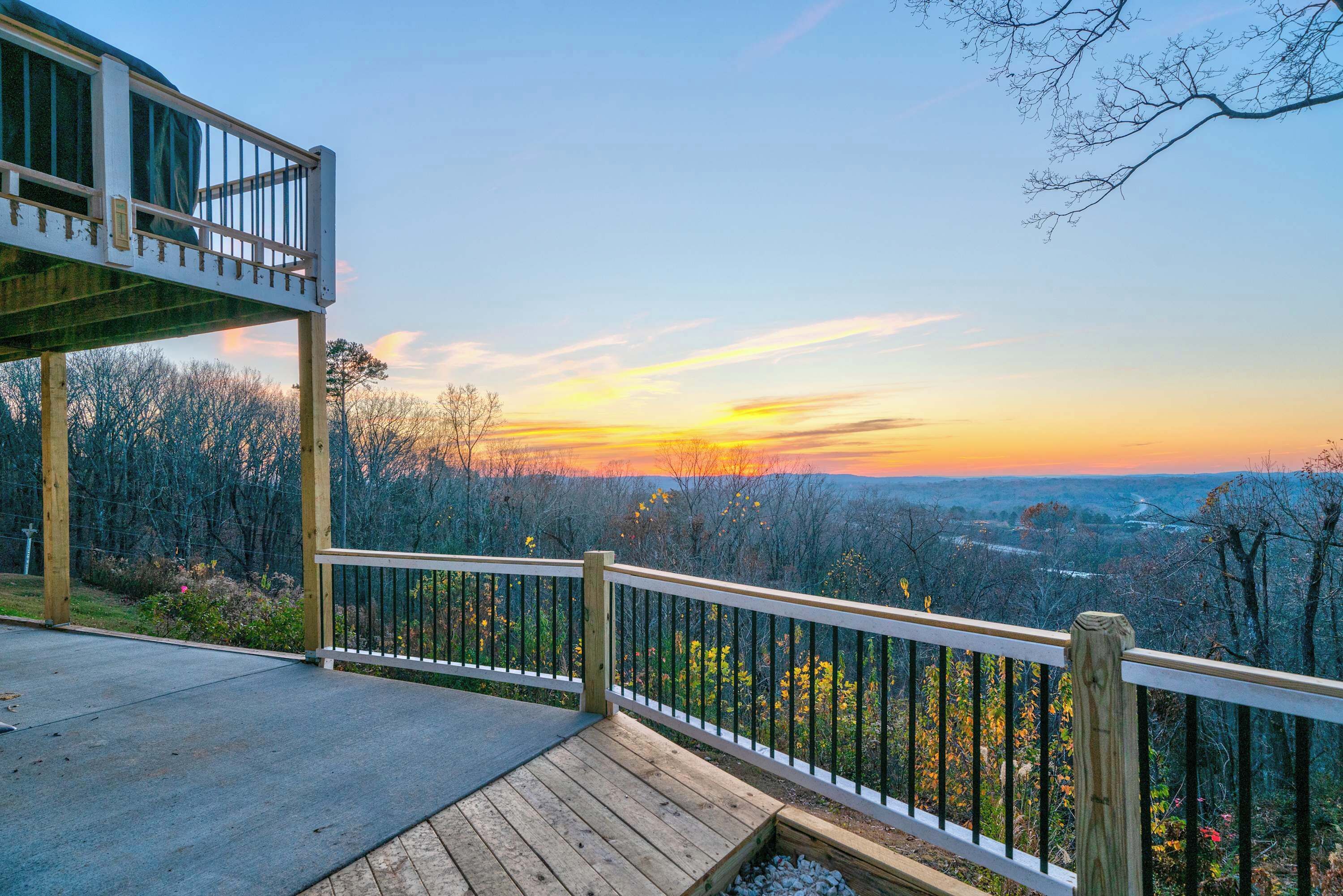 A wooden deck with a railing overlooking a forested landscape during sunset.