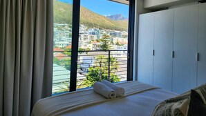 Habitación de hotel con un ventanal grande, una cama con ropa de cama blanca y vistas a la ciudad y montañas.