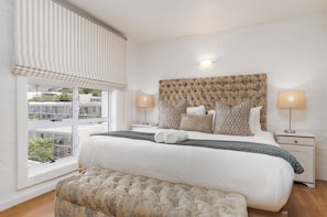 A bedroom with a large bed, two bedside lamps, a window with blinds, and a view of buildings outside.