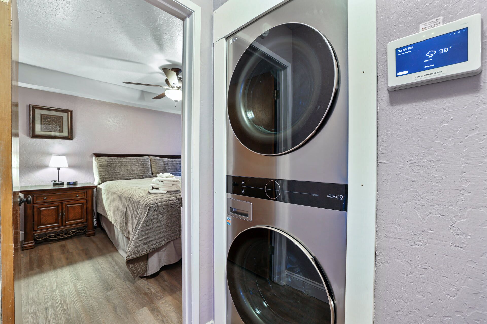 A laundry room with two front-loading washing machines, a bed with a headboard, a nightstand, a lamp, and a ceiling fan.