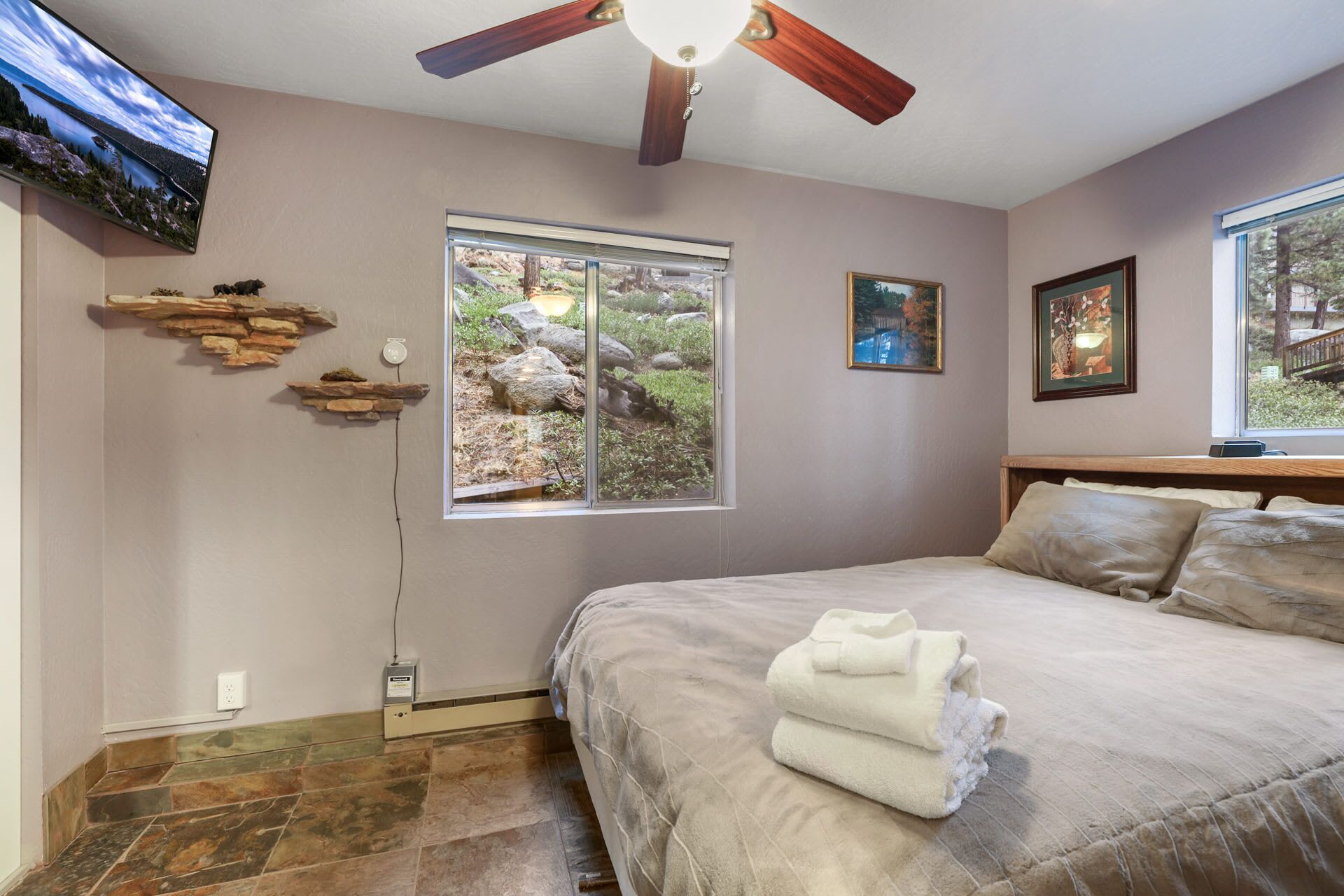 Une chambre à coucher avec un lit, un ventilateur de plafond, un téléviseur fixé au mur et une fenêtre donnant sur des rochers et de la verdure.