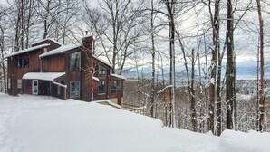 Une cabane en bois, dotée d’un toit à forte pente, entourée d’arbres couverts de neige et avec des montagnes en arrière-plan.