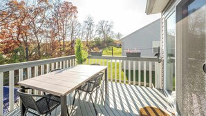 A wooden deck with a table and chairs, a white railing, and a view of trees and a house in the background. - Ark Adventure Retreat (Walton)