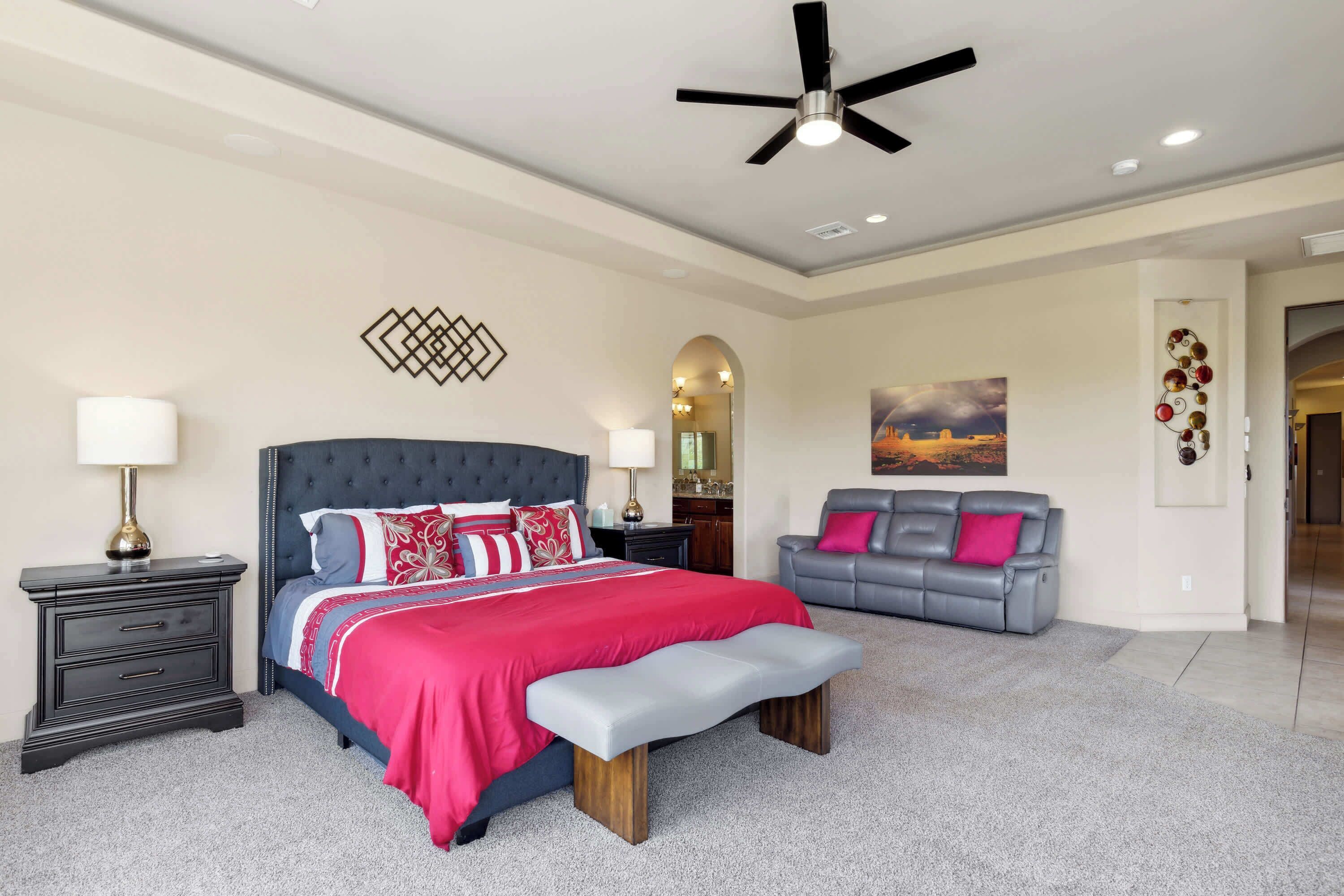 A bedroom with a large bed, a grey sofa, a bench, a ceiling fan, and a painting on the wall.