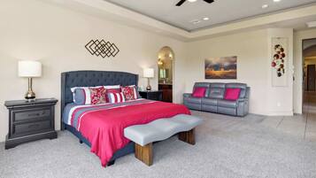 A bedroom with a large bed, a grey sofa, a bench, a ceiling fan, and a painting on the wall.