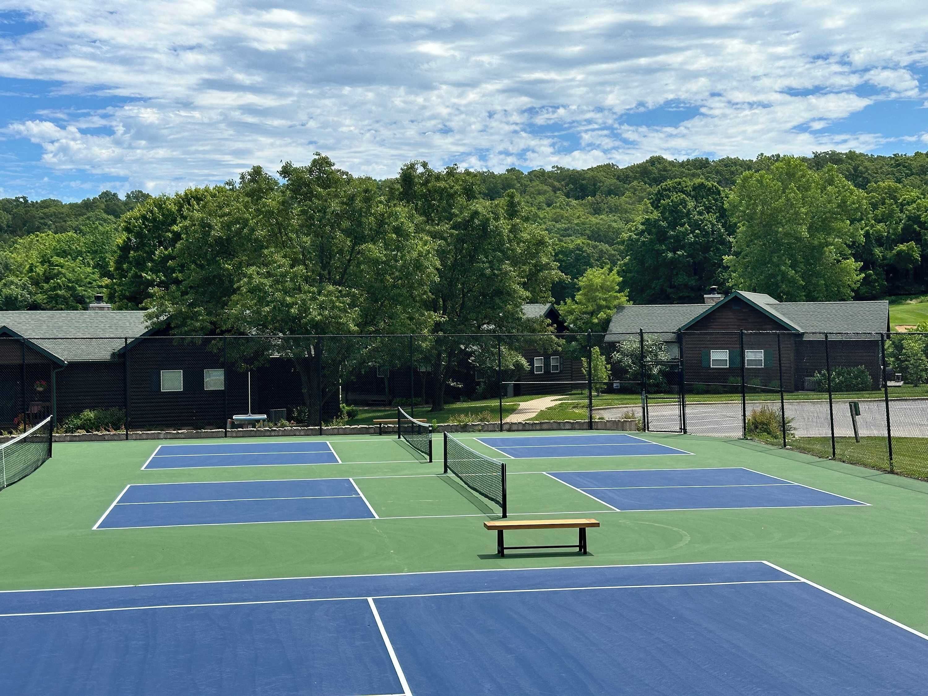 Un court de tennis comprenant plusieurs terrains, entouré d’une clôture, avec des cabanes en bois en arrière-plan.