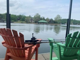 Vista desde una ventana con dos sillas Adirondack, una roja y otra verde, con vista a un lago tranquilo donde se ven una casa y un pabellón al fondo.