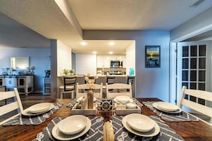 Uma sala de jantar com uma mesa de madeira posta para quatro, uma cozinha ao fundo e um quadro na parede.