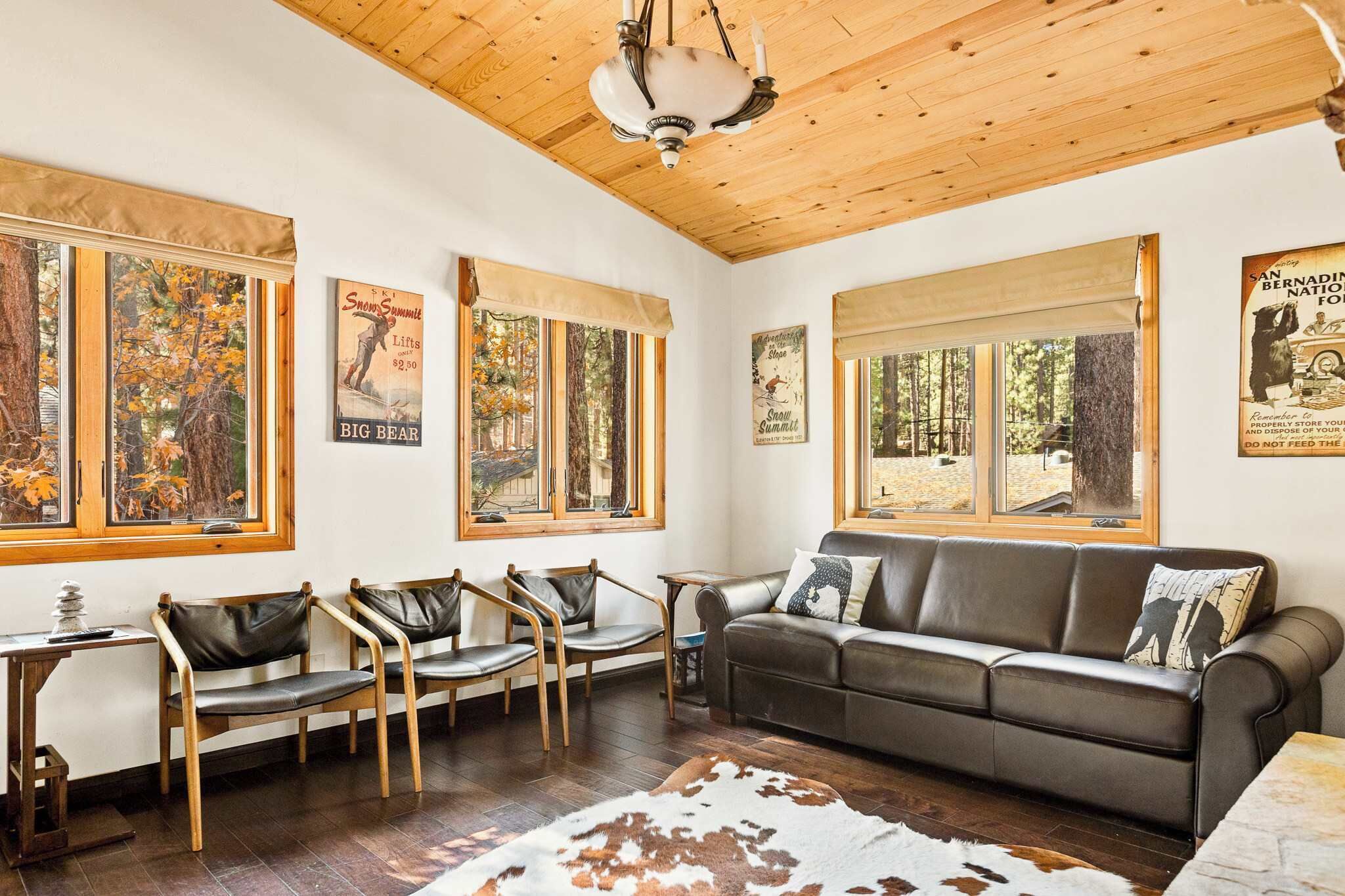 Een gezellige woonkamer met een houten plafond, grote ramen met uitzicht op bomen, een leren bank en drie fauteuils.