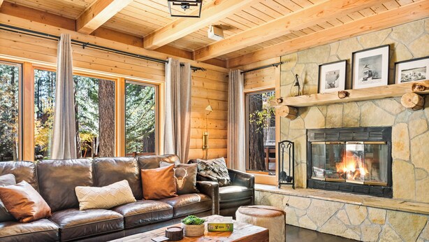A cozy living room with a stone fireplace, leather sofa, and large windows overlooking a forest. - Summit Pine Lodge 4 BDR Minutes From Bear Lake! (Big Bear Lake)