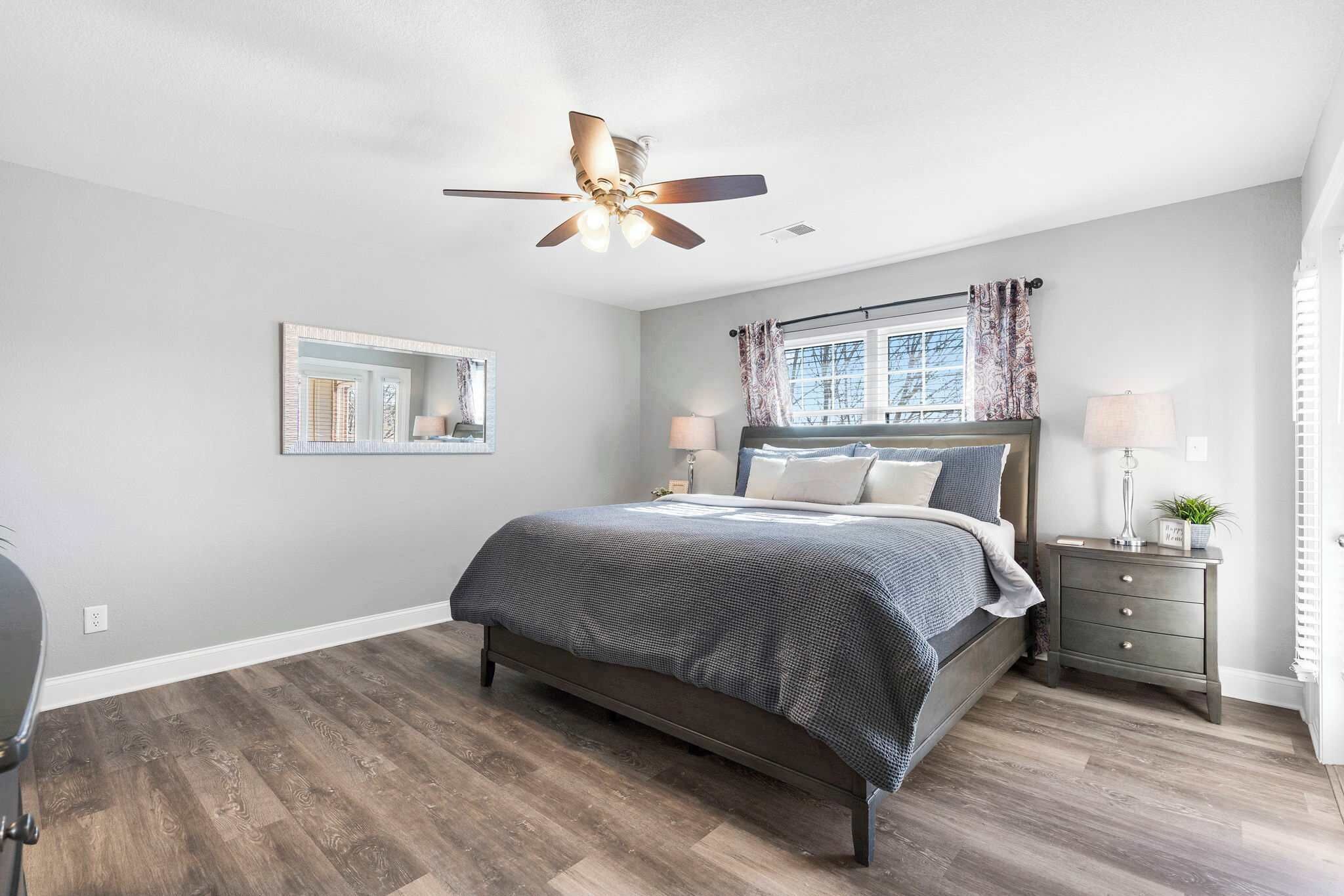 A bedroom with a bed, bedside tables, a ceiling fan, a window with curtains, and a mirror on the wall.