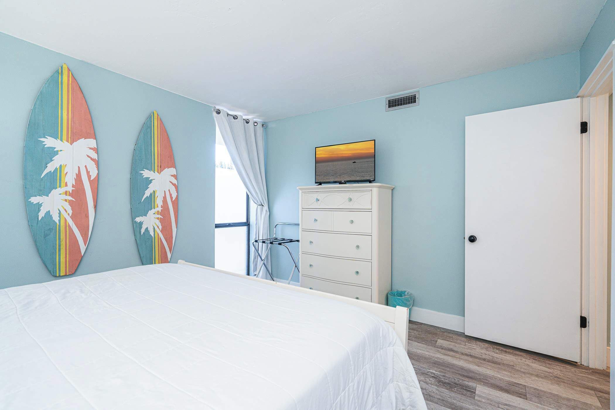 A bedroom with a bed, a dresser, a TV on top of the dresser, and two surfboards on the wall.
