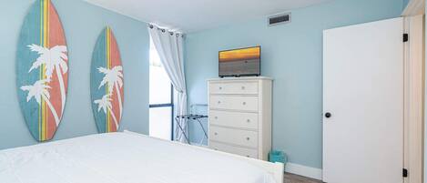 A bedroom with a bed, a dresser, a TV on top of the dresser, and two surfboards on the wall.